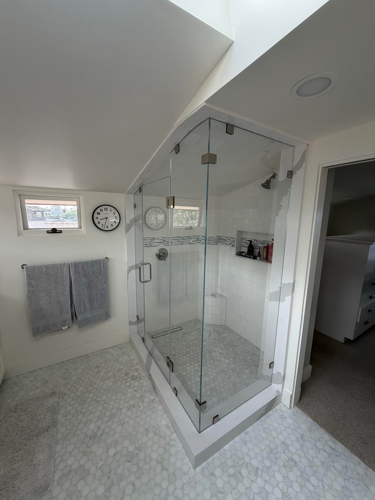 Frameless-glass corner shower under the sloped ceiling with a glass-mosaic accent band and marble-look hex floor